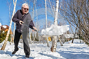 Senior man throwing snow with shovel from private house yard in winter on bright sunny day. Elderly person removing snow in garden