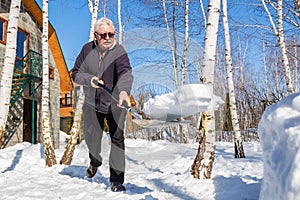 Senior man throwing snow with shovel from private house yard in winter on bright sunny day. Elderly person removing snow in garden