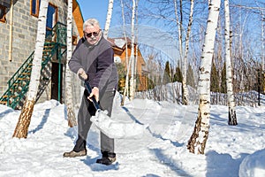 Senior man throwing snow with shovel from private house yard in winter on bright sunny day. Elderly person removing snow in garden