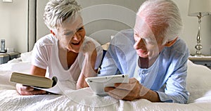 Senior man using digital tablet while woman reading a book on bed