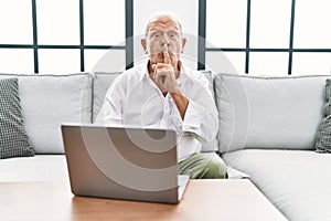 Senior man using laptop at home sitting on the sofa asking to be quiet with finger on lips