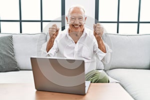 Senior man using laptop at home sitting on the sofa excited for success with arms raised and eyes closed celebrating victory