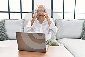 Senior man using laptop at home sitting on the sofa with hand on head for pain in head because stress