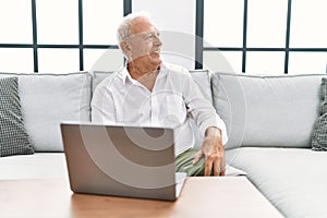 Senior man using laptop at home sitting on the sofa looking away to side with smile on face, natural expression