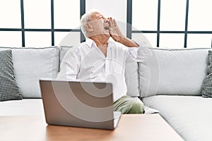 Senior man using laptop at home sitting on the sofa shouting and screaming loud to side with hand on mouth