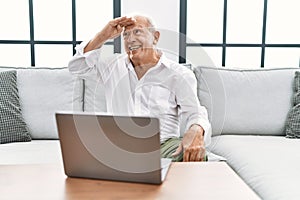 Senior man using laptop at home sitting on the sofa very happy and smiling looking far away with hand over head