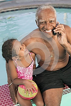 Senior man using mobile phone sitting by pool with granddaughter (5-6) portrait.
