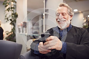 Senior Man Waiting To Have Hair Cut In Hairdressing Salon Looking At Mobile Phone