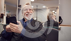 Senior Man Waiting To Have Hair Cut In Hairdressing Salon Looking At Mobile Phone