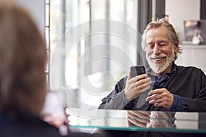 Senior Man Waiting To Have Hair Cut In Hairdressing Salon Looking At Mobile Phone