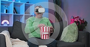 Senior man wearing virtual reality glasses eating popcorn while sitting on sofa at home and watching movie.