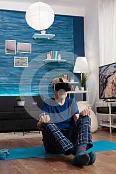 Senior man wearing virtual reality headset meditating while sitting in lotus position on yoga mat