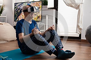 Senior man wearing virtual reality headset meditating while sitting in lotus position on yoga mat