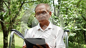 Senior man in wheelchair using digital tablet, looking at screen, reading e-book, chatting online with grandchild in the park.