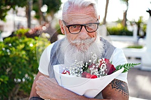 Senior man with white beard and mustache smiling to the camera, waiting for his date with flowers in hands