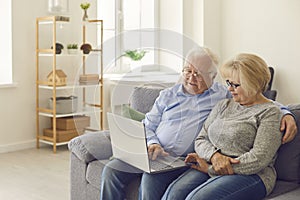 Senior man and woman sitting on sofa in living room watching movie on laptop together.