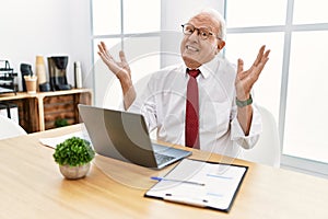 Senior man working at the office using computer laptop clueless and confused expression with arms and hands raised