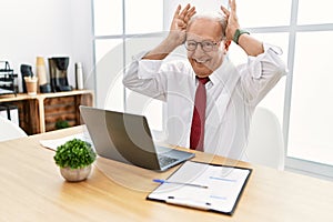 Senior man working at the office using computer laptop doing bunny ears gesture with hands palms looking cynical and skeptical