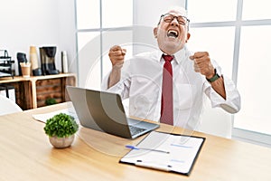 Senior man working at the office using computer laptop excited for success with arms raised and eyes closed celebrating victory