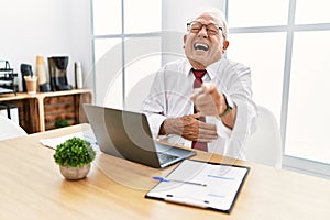 Senior man working at the office using computer laptop laughing at you, pointing finger to the camera with hand over body, shame