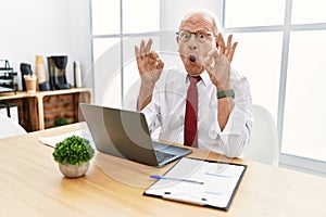Senior man working at the office using computer laptop looking surprised and shocked doing ok approval symbol with fingers