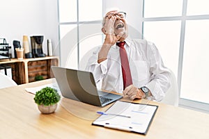 Senior man working at the office using computer laptop shouting and screaming loud to side with hand on mouth