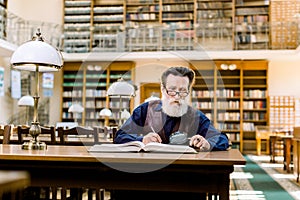 Senior man working in old library, sitting at the table with books and magnifying glass, making notes. Student old man