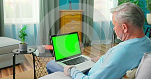 Senior Man Works on a Laptop Computer with Green Mock-up Screen Sitting on a Couch