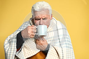 Senior man wrapped in blanket drinking hot beverage on yellow background. Cold symptoms
