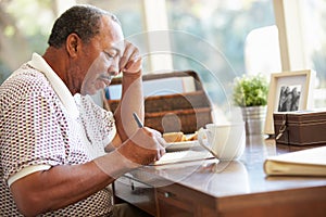 Senior Man Writing Memoirs In Book Sitting At Desk