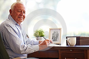 Senior Man Writing Memoirs In Book Sitting At Desk