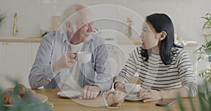 Senior man and young woman drinking tea and talking enjoying meal and conversation in kitchen at home