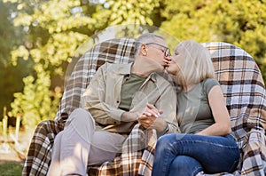 Senior married couple resting outdoors and kissing, enjoying holiday while sitting on wicker chairs in garden