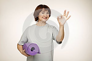 Senior mature pretty woman holding yoga mat doing ok sign with fingers, smiling friendly gesturing excellent symbol
