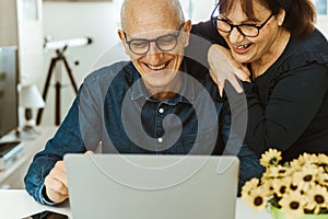 Senior middle aged happy couple embracing using laptop together - smiling elderly family reading the news and shopping online from