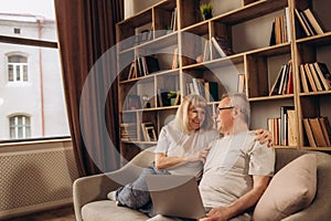 Senior middle aged happy couple embracing using laptop together, smiling elderly family reading news, shopping online at