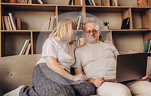 Senior middle aged happy couple embracing using laptop together, smiling elderly family reading news, shopping online at