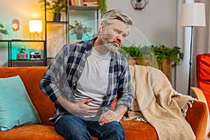 Senior middle-aged man sits on couch feeling sudden strong abdominal stomach ache gastritis problem