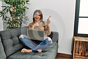 Senior middle east woman using laptop sitting on the sofa at home smiling and looking at the camera pointing with two hands and