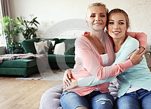 Senior mom and her adult daughter are hugging, looking at camera and smiling. At Home.