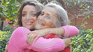 Senior mother with gray hair with her adult daughter are hugging each other during sunny day outdoors in the garden