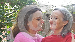 Senior mother with gray hair with her adult daughter are hugging each other during sunny day outdoors in the garden