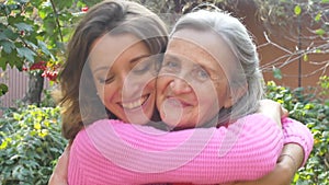 Senior mother with gray hair with her adult daughter looking at the camera in the garden and hugging each other during