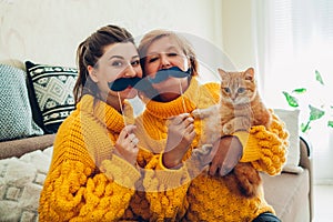 Senior mother and her adult daughter taking selfie with cat using photo booth props at home. Mother`s day concept