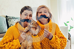 Senior mother and her adult daughter taking selfie with cat using photo booth props at home. Mother`s day concept