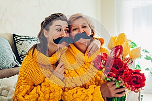 Senior mother and her adult daughter taking selfie with flowers using photo booth props at home. Mother`s day concept