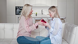 Senior mother presenting birthday gift box to her happy adult daughter smiling bonding embracing, mature woman