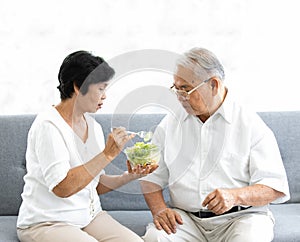 Senior old Asian man and woman lovers eating vegetables salad together on sofa in home