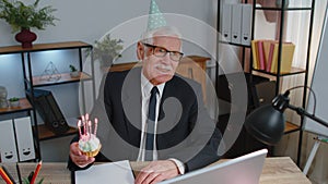 Senior old business man celebrating birthday in office holding small cake with candles making a wish