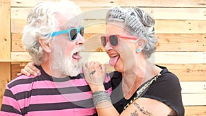 Senior old caucasian couple with alternative look and cheerful lifestyle engage and have fun in camera portrait with wooden backgr